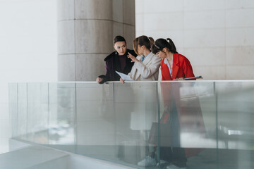 Three professionally dressed women engage in discussion at a contemporary workplace, suggesting...