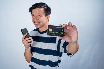 Asian man in striped shirt holding a smartphone in one hand and showing a credit card toward the camera with a surprised expression, against a clean studio background