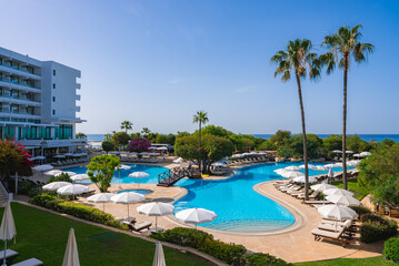 A resort in Cyprus features a curving pool with sun loungers, white umbrellas, a modern hotel, palm trees, and a glimpse of the Mediterranean Sea.