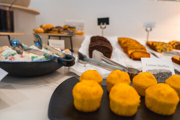 A dessert buffet featuring lemon muffins, Turkish delights with tongs, chocolate cake slices, and assorted pastries in a hotel or resort setting.
