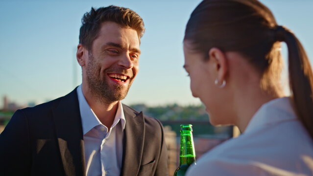 Laughing coworkers clinking beer bottles spending sunset at rooftop closeup.