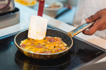 A person cooks an omelet with cheese, ham, and mushrooms in a frying pan on an induction stove. Blurred kitchen elements are visible in the background.