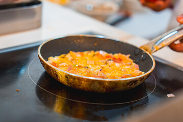 A frying pan on a modern induction stove holds a vibrant mix of diced tomatoes, onions, and eggs. The background suggests a kitchen setting.