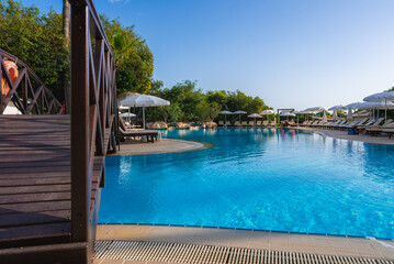 A serene outdoor pool surrounded by sun loungers, umbrellas, and lush greenery. A wooden bridge adds charm, set under clear blue skies in Cyprus.