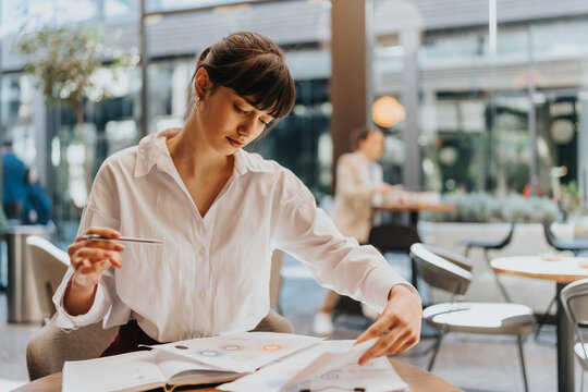 A focused young woman reviews paperwork, analyzing diagrams and data at a table within a contemporary cafe environment.