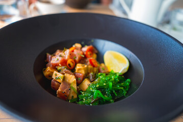 A black bowl features seaweed salad, marinated fish or tofu, red chili slices, and a lemon wedge. The blurred background suggests a dining setting.