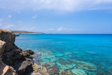 Crystal clear turquoise waters meet rocky formations along the Cyprus coast. A distant stretch of land with urban development is visible under a blue sky.