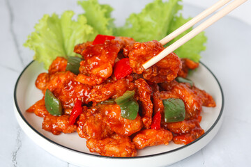 Person using chopsticks to eat homemade sweet and sour fish with bell peppers, served on a white plate with fresh lettuce on the side. Captured with natural light on marble background.