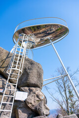Visitors admire the panoramic views from Sipka lookout platform in the Jizera Mountains, with a bright blue sky and rocky terrain surrounding the structure, offering a unique vantage point.