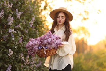 Beautiful woman with basket of lilac flowers near bush outdoors in morning