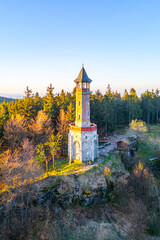 The Stepanka Lookout Tower stands tall against a clear spring sky in Korenov, surrounded by lush greenery. Sunlight illuminates the tower, highlighting its architectural beauty.