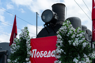 Historic steam locomotive on display at a local celebration under a clear sky with floral decorations and red flags in the background