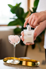 A cotton candy cocktail in a glass, with pink fluffy candy dissolving into the drink. A hand sprays perfume labeled "LOVE IS... MADE FOR YOU" over it.