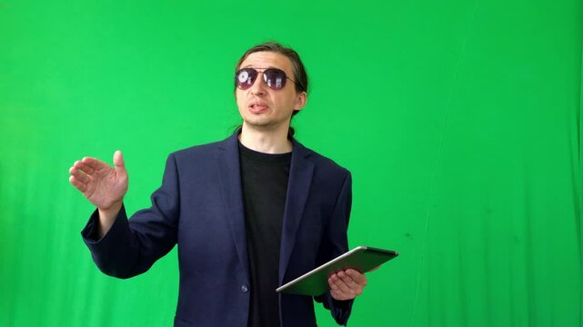 Authoritative and engaging mood as a man dressed as a business coach, wearing sunglasses, presents or explains while holding a tablet against a vibrant green screen ideal for corporate or educational