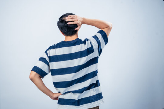Asian man in navy and white striped t-shirt seen from behind, scratching head with one hand and resting the other on hip, standing against plain light background - Powered by Adobe