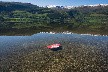 Norwegian heart: a heart-shaped Norwegian flag in a clear lake against a backdrop of mountains and a summer landscape.