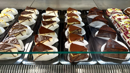 A variety of delicious cakes arranged in rows behind a glass display case in a bakery, including chocolate-glazed, cocoa-dusted, and berry cream slices, each served in white paper cups, sweets concept