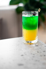 A vibrant layered cocktail in a tall glass, featuring green and yellow liquids, garnished with a cherry on a skewer. Set on a speckled countertop with a blurred background, evoking a refreshing 