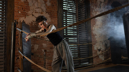 Fototapeta premium A woman in a boxing gym, portrait by the ropes, cinematic style, dark tones. Training and overcoming oneself, motivation