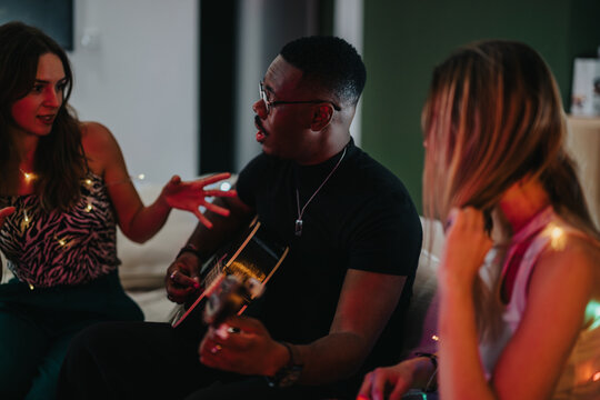 A diverse group of friends gather in a cozy room, sharing moments of music and joy as one plays the guitar and others join in conversation, celebrating companionship and creativity.