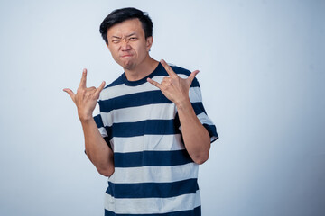 Asian man in striped shirt making a rock and roll hand gesture with both hands, scrunching his face into a fierce expression against a plain light background