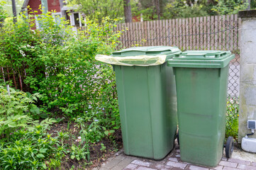 Two green plastic garbage bins stand near fence on private plot, one of them with yellow bag for collecting packaging