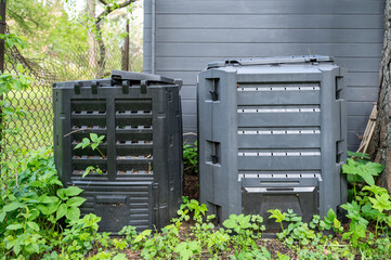 Two plastic compost bins for organic waste stand behind house by wall amidst grass