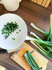 Top view of table surface with cutting board, green onion and knife. Part of onion already cut and lies in bowl nearby