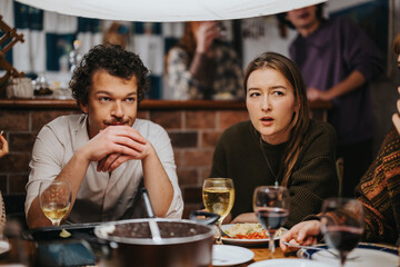 Two young adults are seated indoors at a bar table, enjoying conversation and drinks in a casual setting, surrounded by friends, creating a warm and relaxed social atmosphere.