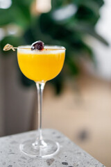 A close-up of a yellow cocktail in a coupe glass, garnished with a dark cherry on a skewer, placed on a terrazzo countertop with a blurred green background, creating a fresh and elegant atmosphere