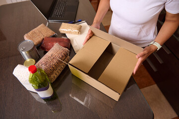 Person Packing Essential Food Items into a Cardboard Box on a Table with Supplies