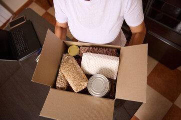 Person Holding a Box Containing Non-Perishable Food Items and Essential Supplies on a Table