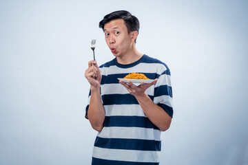 Asian man in striped shirt holds a plate of instant noodles and a fork while smiling...