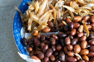 A close-up view of a traditional Malaysian side dish featuring crispy fried anchovies (ikan bilis) and roasted peanuts served on a blue plate. 