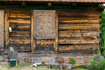 old wooden cabin wall with a boarded-up window and door. Weathered wood planks, stone foundation, and signs of decay are visible. A green bucket and discarded objects lie near the base