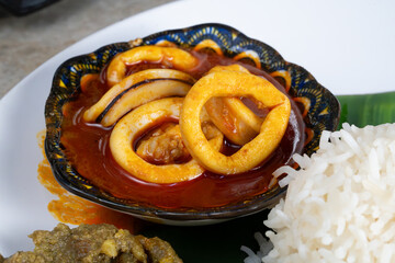 A traditional Malaysian sambal sotong dish, featuring squid rings cooked in spicy chili paste, is served in a small ornate bowl alongside steamed white rice and accompaniments. 