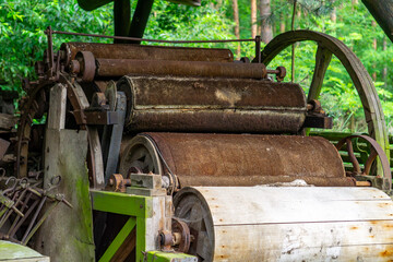 Vintage agricultural machine with large rusty rollers and pulley wheels, standing outdoors surrounded by greenery. The equipment is covered with moss and signs of corrosion.