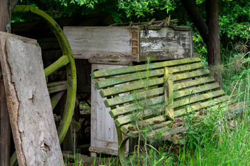 Naklejka premium Old wooden bench covered with moss, standing under a shelter amid tall grass. Next to it are weathered boards and part of a wooden wheel. A forgotten rural corner surrounded by nature.