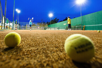 bolas de tenis amarelas em primeiro plano uma quadra ao entardecer com pessoas ao fundo praticando...