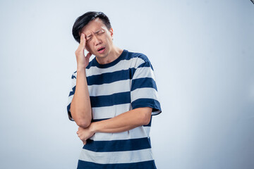 Asian man in a striped shirt touches his forehead and squints his eyes in discomfort, expressing a headache or stress, against a plain light background