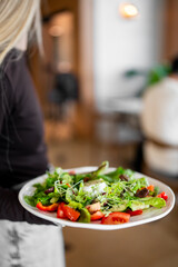 A person holds a plate of fresh salad with vibrant vegetables like tomatoes, lettuce, and microgreens in a restaurant setting. The dish looks colorful, healthy, and appetizing.