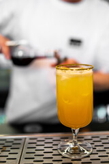 A bartender in a white shirt pours a dark liquid into a glass with a vibrant yellow drink on a bar counter. The focus is on the drink in the foreground, highlighting its color and presentation
