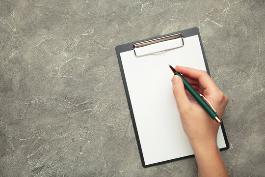 A female hand hold a pen and black clipboard with paper on grey concrete background.