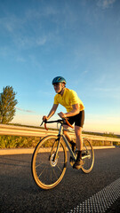 Fototapeta premium Focused cyclist sprints through final stretch of training route at golden hour with low sunlight reflecting off asphalt and sky turning orange. Concept of sport, cycling, racing, training, endurance