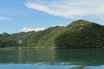 View of the sky, mountains, and sea during the summer afternoon. The sky is scattered with white clouds. The mountains are covered with green trees, the sea is calm. Natural beauty.
