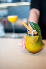 A vibrant tropical cocktail garnished with a cinnamon stick, green leaf, and passion fruit slice, held by a person in a bar setting. The blurred background adds depth and atmosphere.