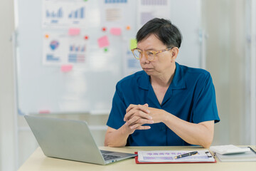 Fototapeta premium Asian man sitting at desk in front of laptop at home preparing budget and financial documents related to household expenses.
