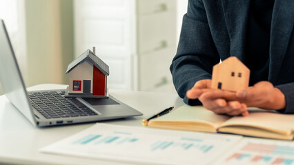 Real estate agent using laptop at desk with miniature model of house, representing real estate investment, buying a house, close up.