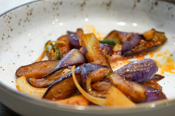 Spicy stir-fried eggplant with onions and peppers served in a white ceramic bowl.  Close up.