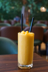 A vibrant mango smoothie in a tall glass with straws, placed on a wooden table, set against a blurred cafe background.